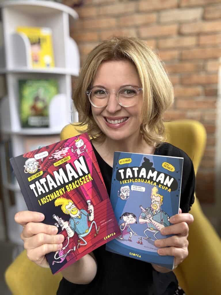 Kobieta trzyma dwie książki z serii Tataman, uśmiechnięta, w tle biblioteka.