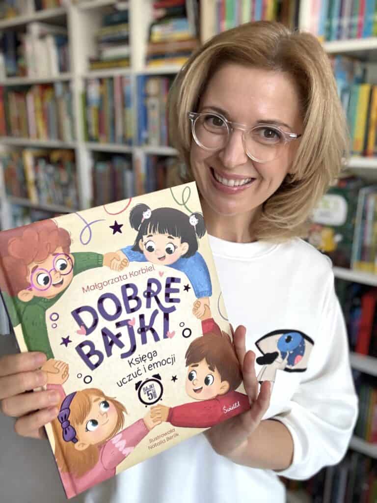 Uśmiechnięta kobieta w okularach i białej bluzie trzyma książkę „Dobre bajki. Księga uczuć i emocji” Małgorzaty Korbiel, na tle regałów z książkami