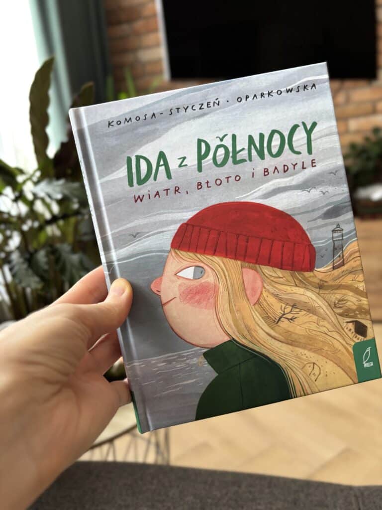 Okładka książki „Ida z Północy. Wiatr, błoto i badyle” z rysunkiem dziewczynki w czerwonej czapce i zielonym płaszczu na wietrznym wybrzeżu