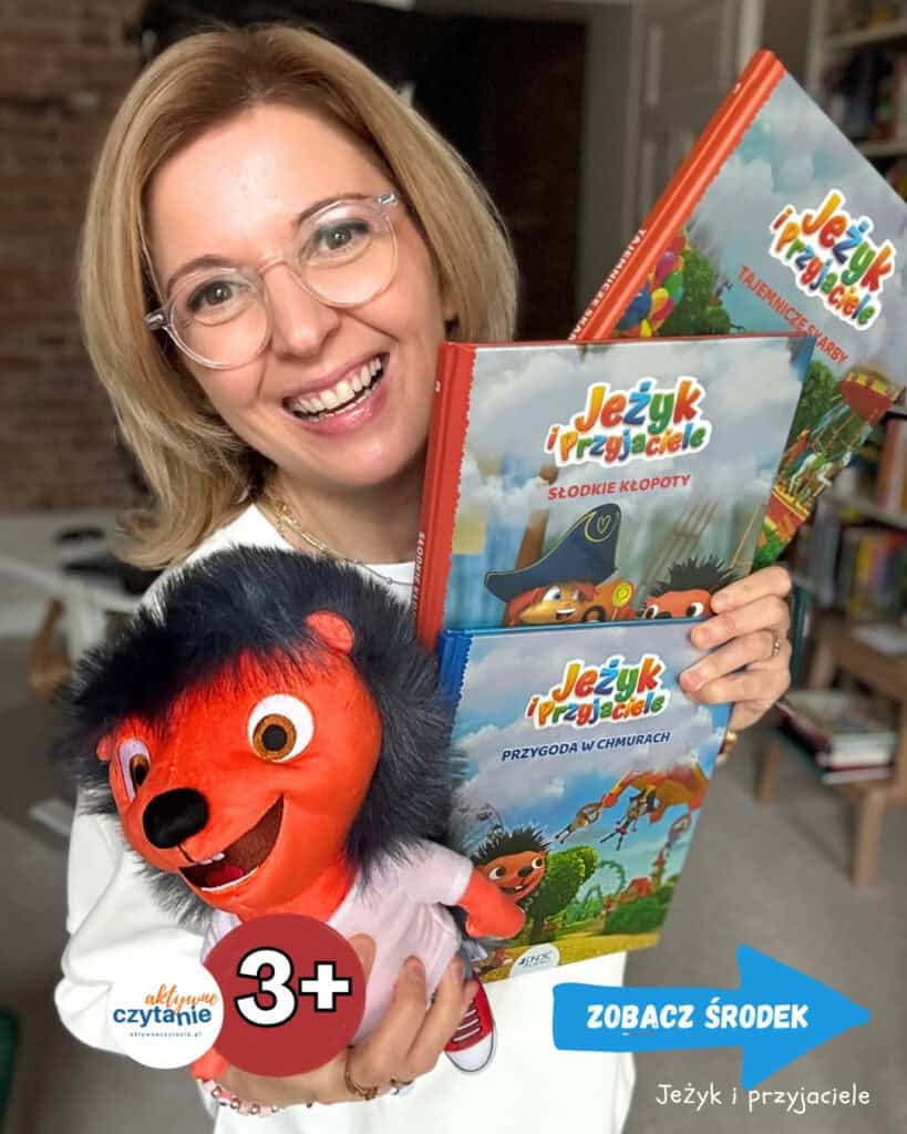 Uśmiechnięta kobieta w okularach trzyma trzy książki z serii „Jeżyk i Przyjaciele” oraz pluszową maskotkę czerwonego jeża