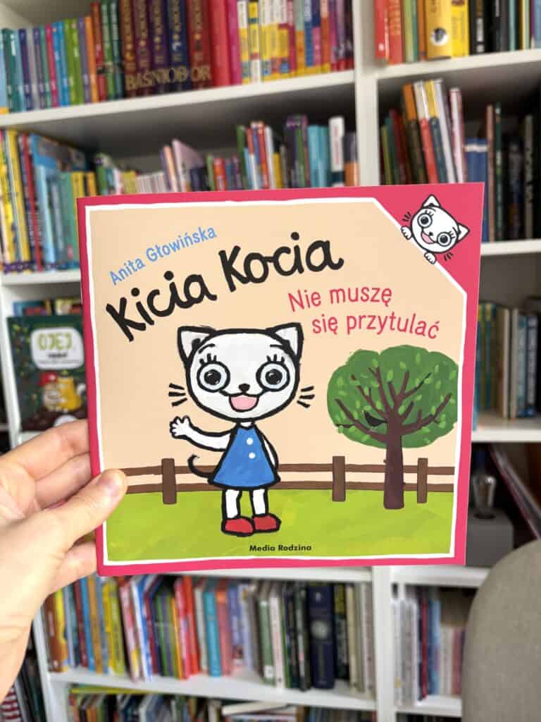 Okładka książki „Kicia Kocia. Nie muszę się przytulać” Anity Głowińskiej: biała kotka w niebieskiej sukience machająca ręką obok drzewa i płotu Count: roughly 150 characters, slightly over 120. Let me shorten. Okładka książki „Kicia Kocia. Nie muszę