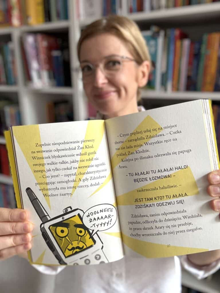 Kobieta w okularach trzyma otwartą książkę z żółtymi ilustracjami, na której walkie-talkie krzyczy „WOOLNEEE ŻAAAAR-TYYYY!”, a strona zawiera napis „TU AŁAŁA!