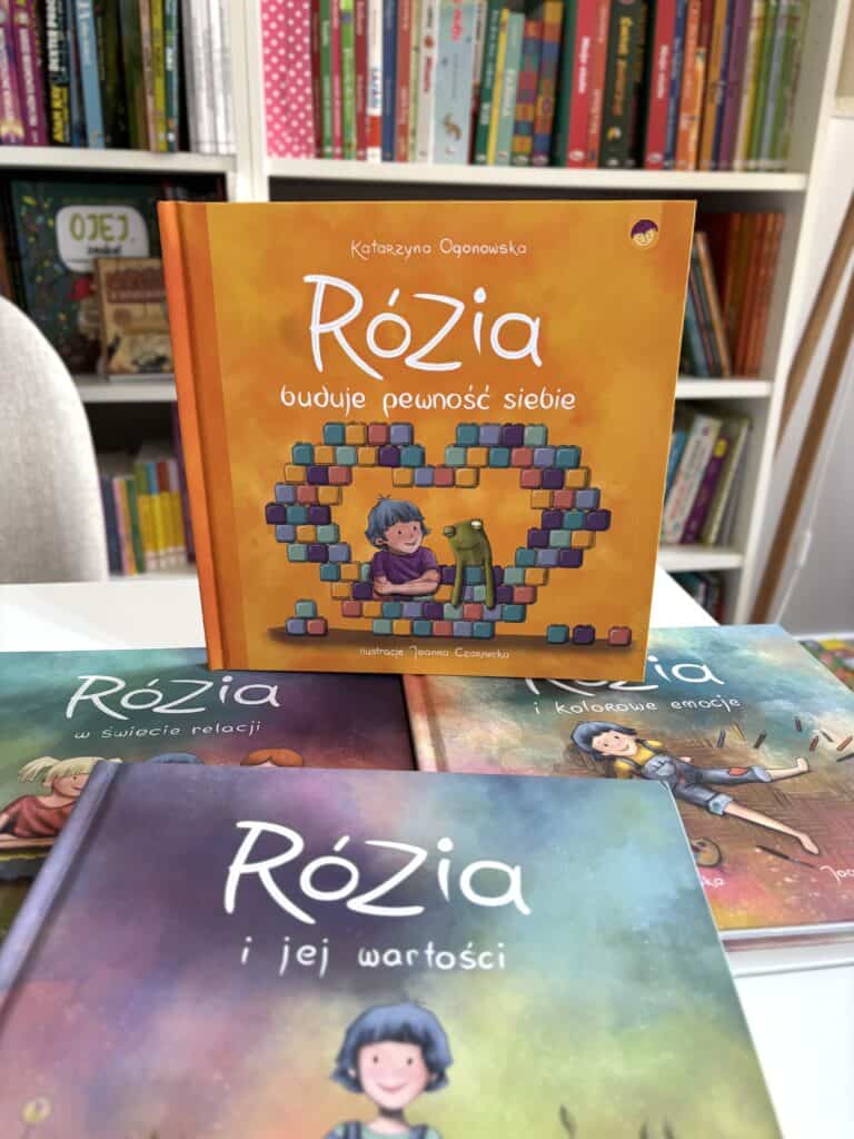 Książki o Rózi w twardych okładkach na białym stole, na przodzie tytuł „Rózia buduje pewność siebie” z ilustracją dziewczynki i zielonego potwora z klocków Hmm, I should keep it under 120 characters and one sentence, no period at end. Let me craft a