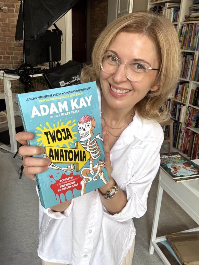 Uśmiechnięta kobieta w białej koszuli i okularach trzyma książkę „Twoja anatomia” Adama Kaya, z ilustracją szkieletu z mózgiem