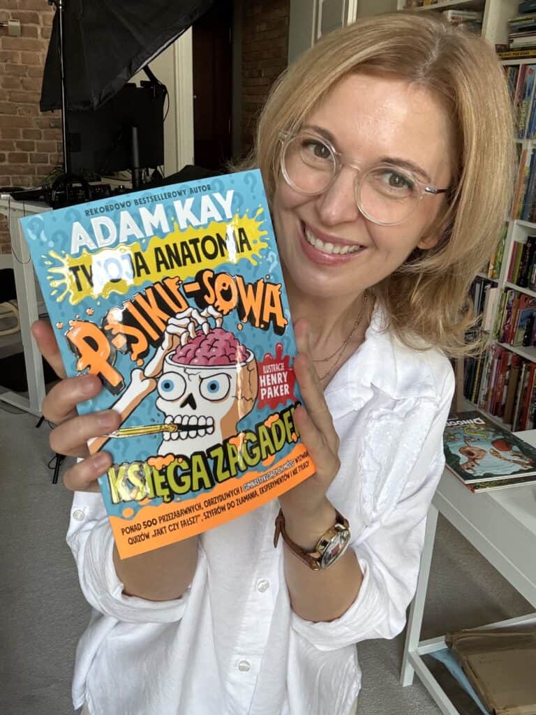 Uśmiechnięta kobieta w białej koszuli i okularach trzyma książkę Adam Kay „Twoja anatomia. Psikusowa księga zagadek”, ilustrowaną przez Henry'ego Pakera